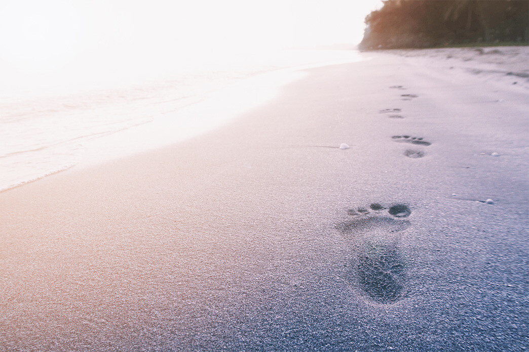 Footprints in the sand leading towards the ocean at sunrise or sunset, with a serene beach and gentle waves.